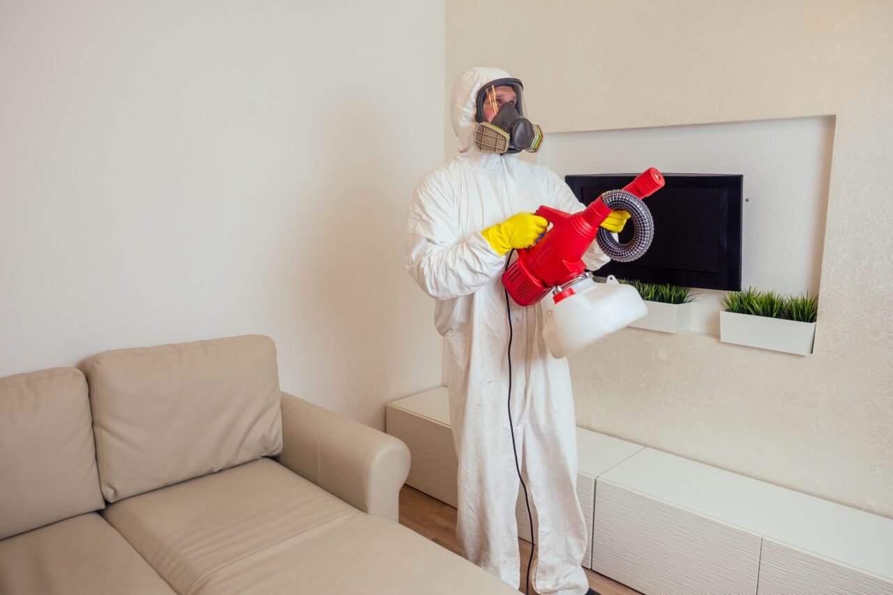pest control worker in uniform spraying pesticides under couch in living lounge room