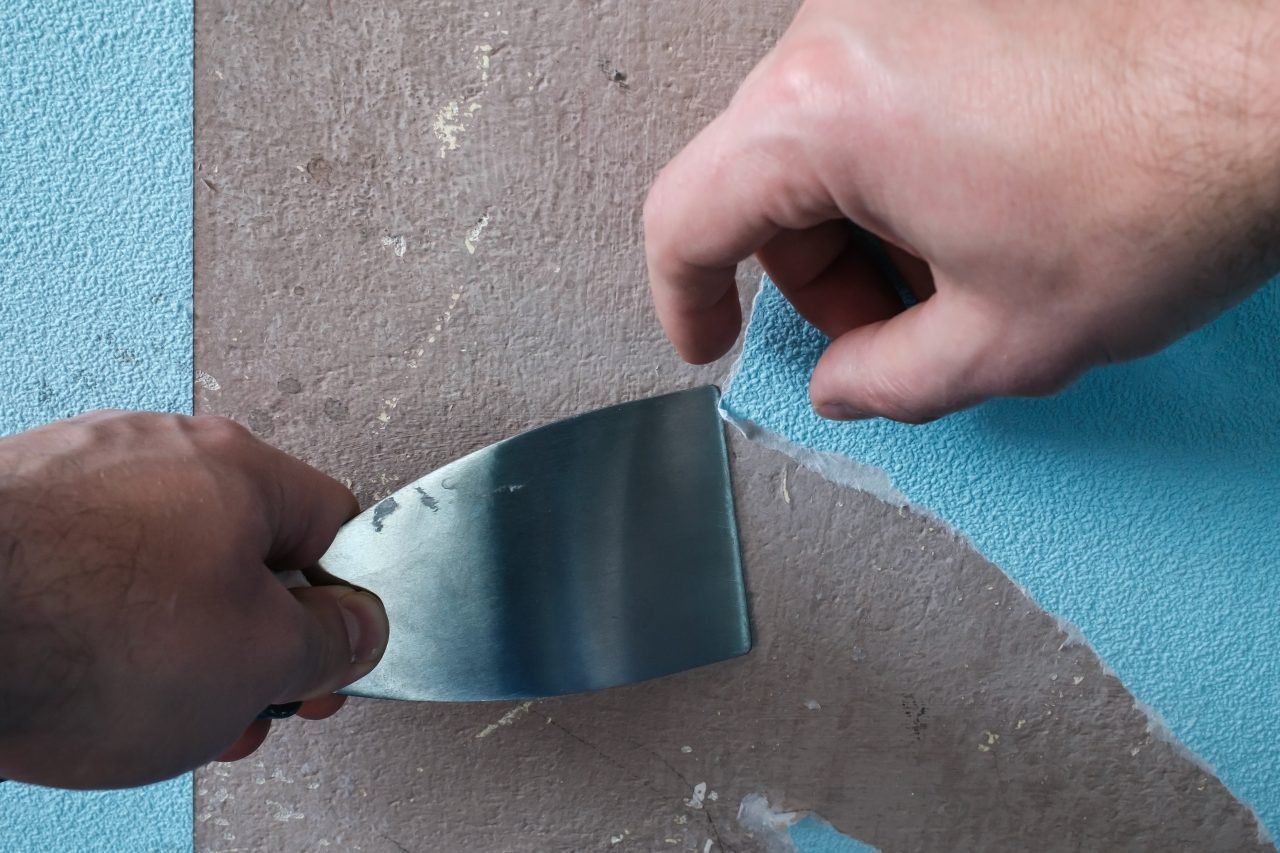Hand removes old blue wallpaper from the walls, using a spatula. House renovation.