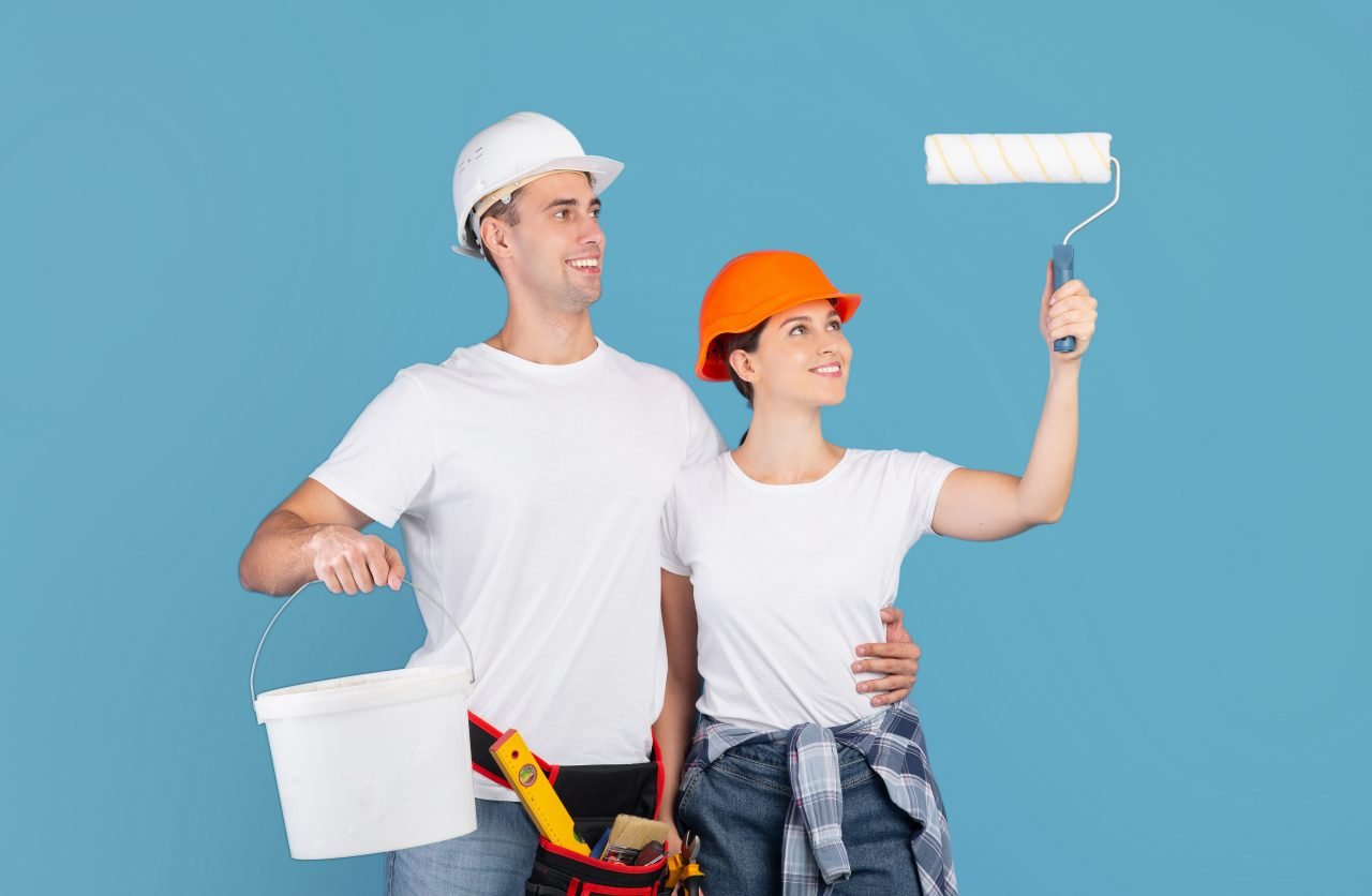 House Renovation Services. Happy Couple In Hardhats Holding Paint Roller And Bucket