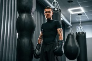 Standing and looking at the camera. Young tattooed man is practicing boxing indoors in the gym